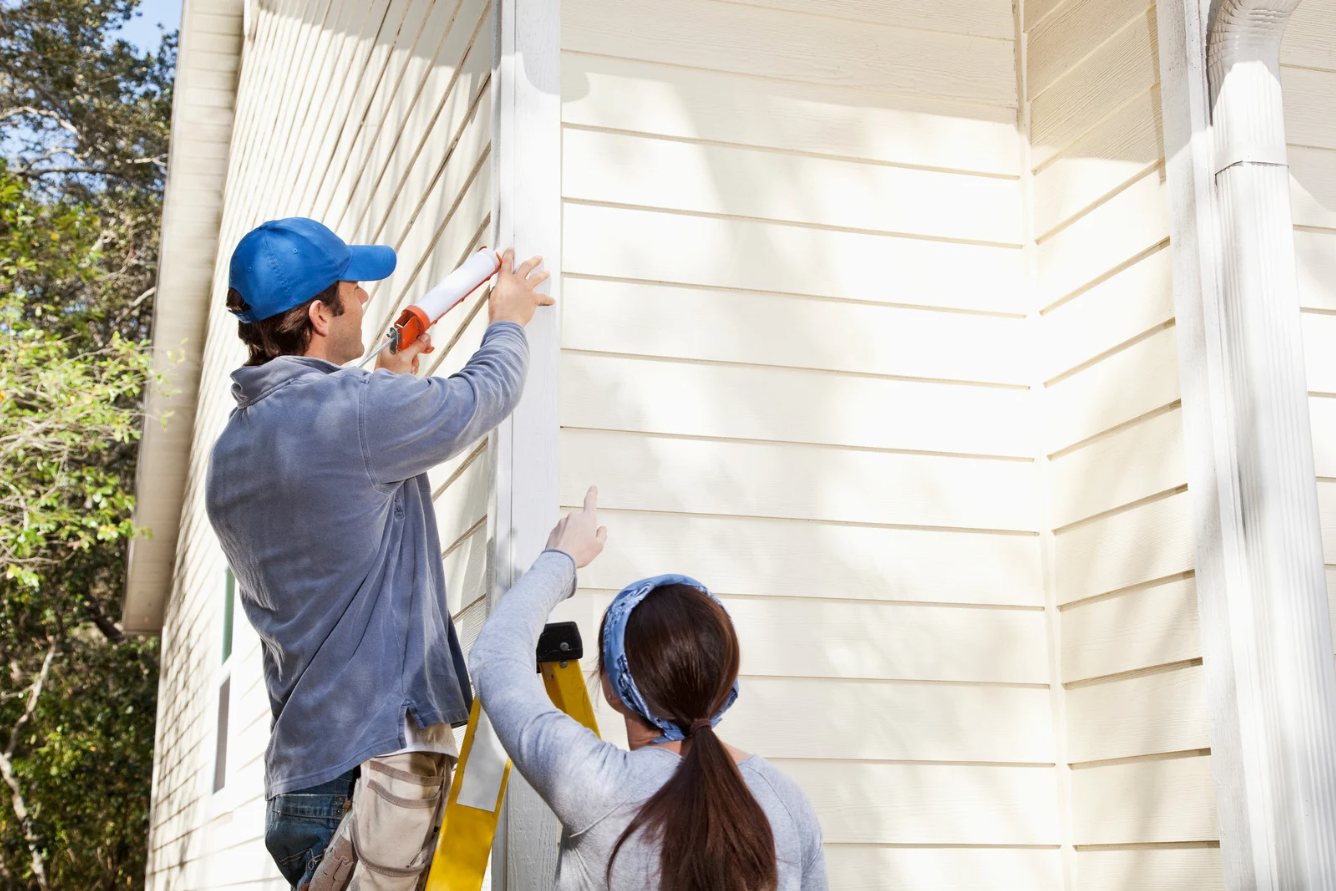 Home repairs outside with man standing on ladder using caulk gun.