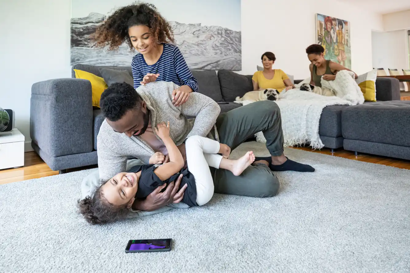 A father plays with two daughters on the living room floor, while their elder sister and mom sit together on the sofa with two dogs.