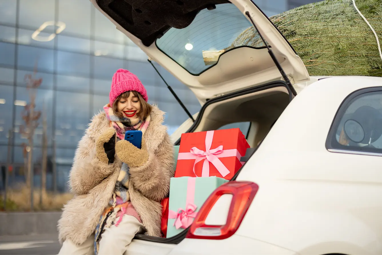 Woman piles Christmas gifts into the trunk of her car as she checks her mobile phone.