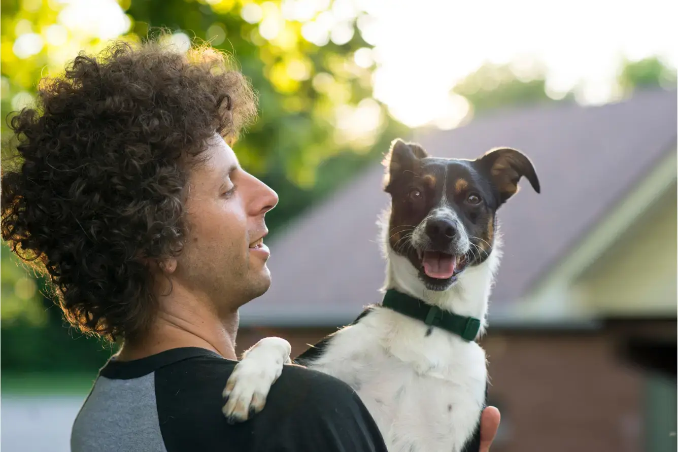Young man with curly hair is holding his dog up in the forest at sunset and looking at her lovingly. Get pet insurance in ontario with firstontario insurance brokers.