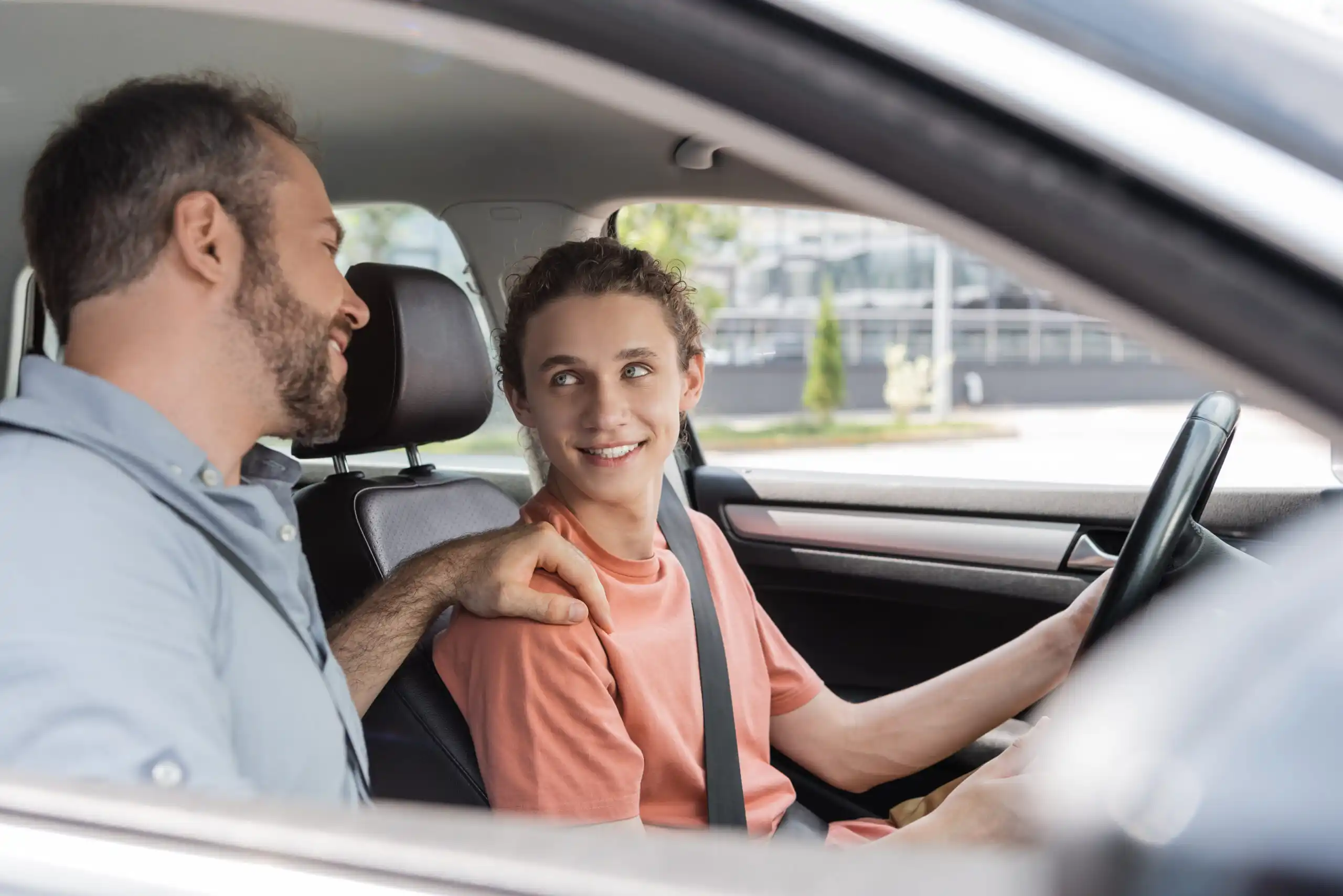 A cheerful father placing a hand on his teenage son's shoulder while teaching him how to drive, symbolizing guidance and support for young drivers seeking car insurance.