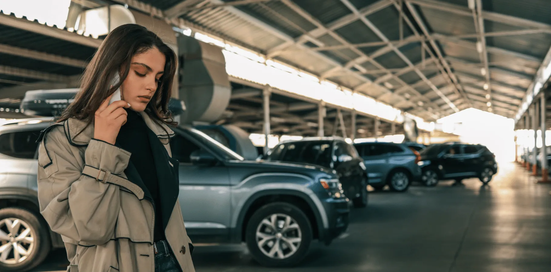 Vehicle owner standing in a covered parking garage beside parked cars, checking a phone after discovering a missing vehicle.